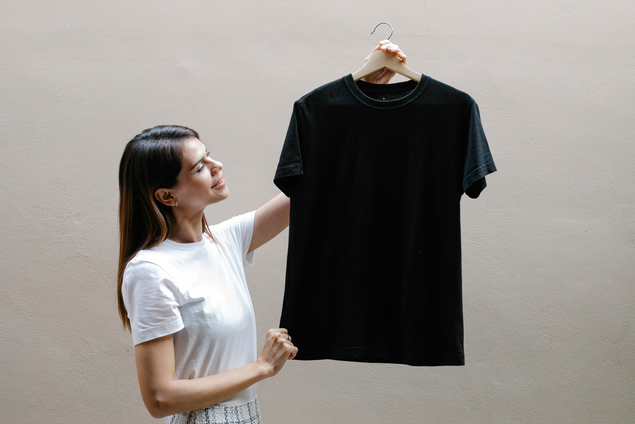 pexels photo 7319141 7319141 Side view of self assured young woman with long dark hair in stylish outfit smiling while holding hanger with black t shirt against gray wall